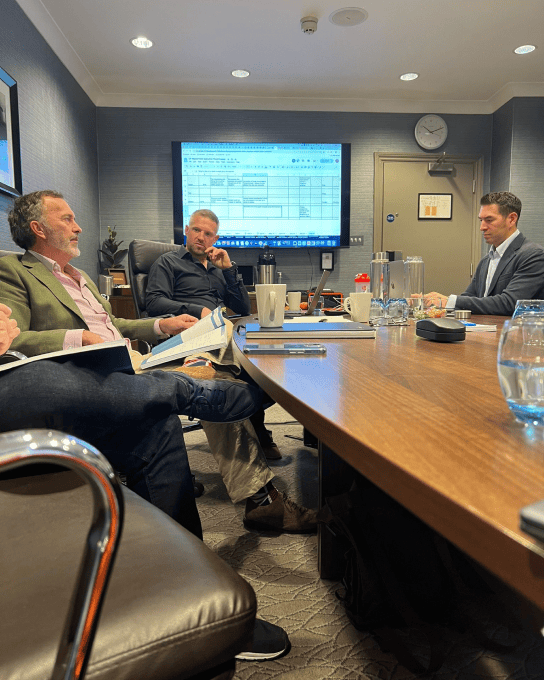 Three men in business attire engaged in a meeting around a conference table with a screen displaying a spreadsheet in the background.