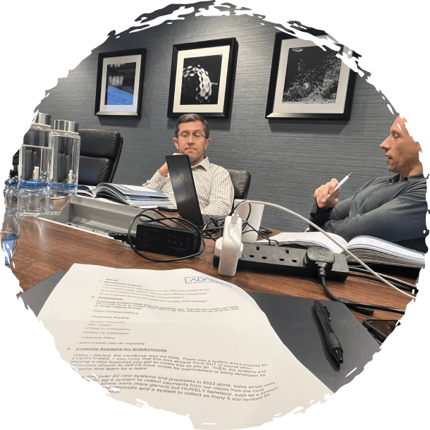 Two men seated at a conference table with laptops, papers, and water bottles during a meeting in a modern office.