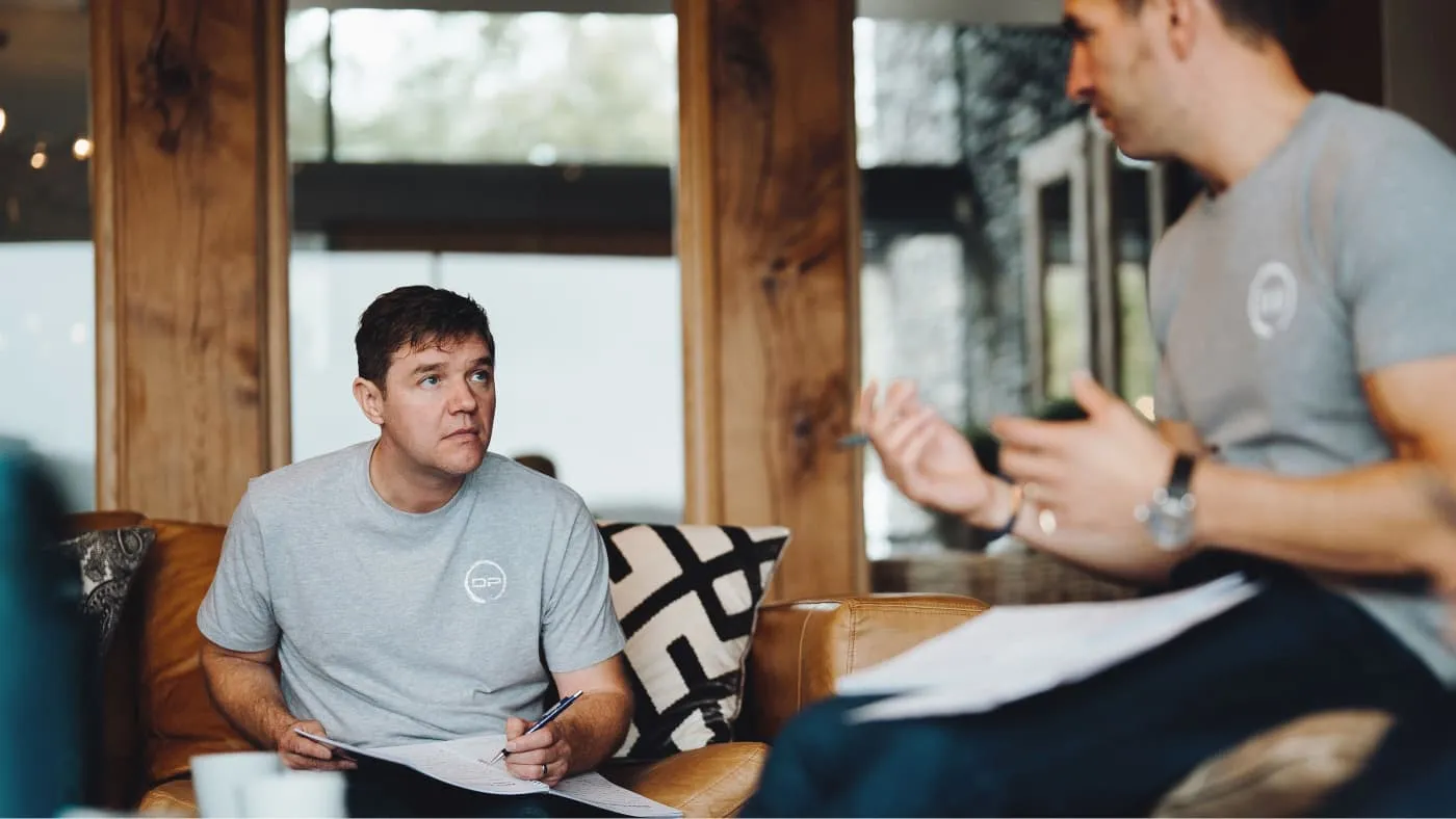 Two men in gray t-shirts having a serious discussion indoors while holding documents and pens.