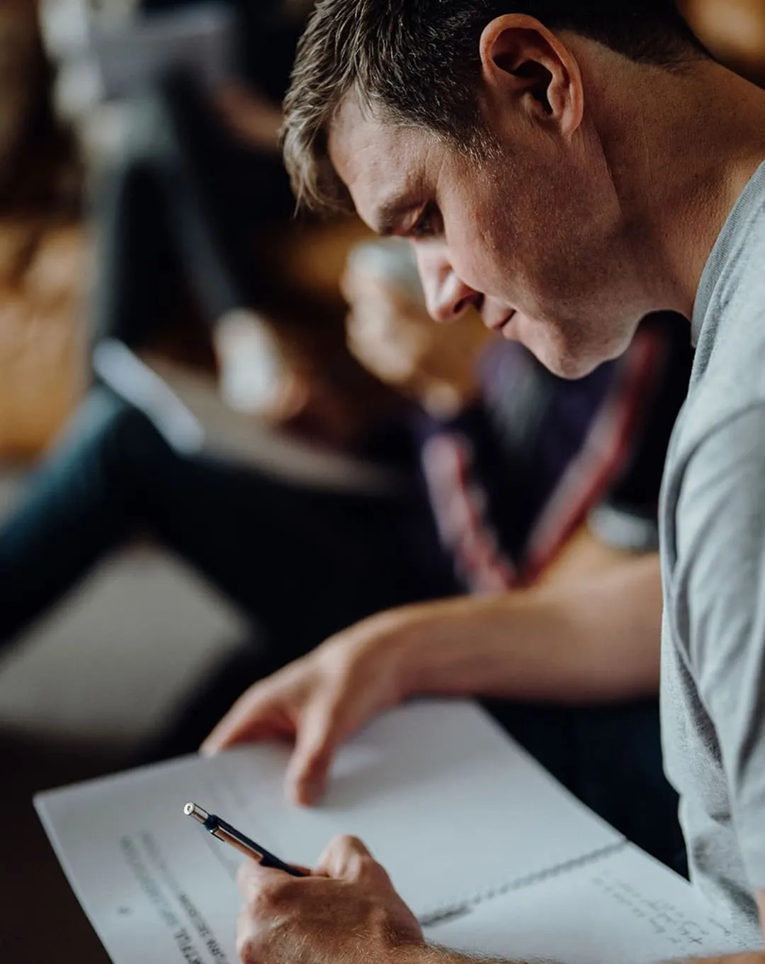 Man leaning forward and writing in a spiral notebook with a pen, with blurred people in the background.