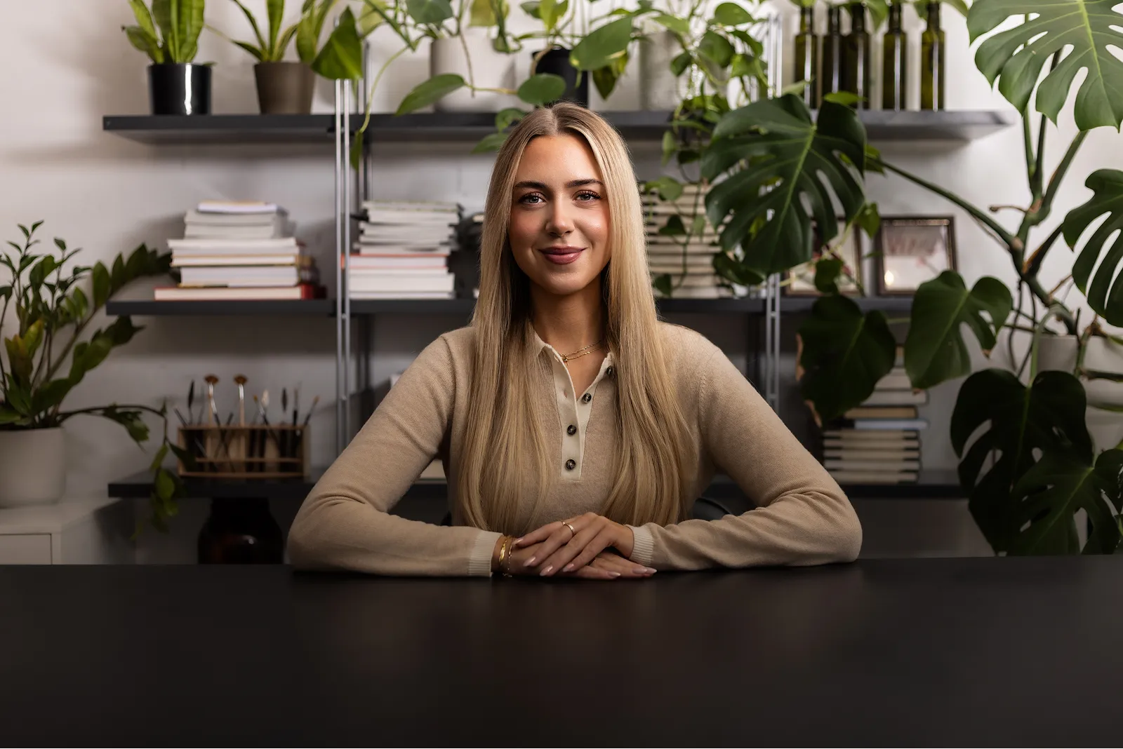 Ung kona með ljóst langt hári sitjandi við borð, bak við hana eru bókahillur og plöntur.