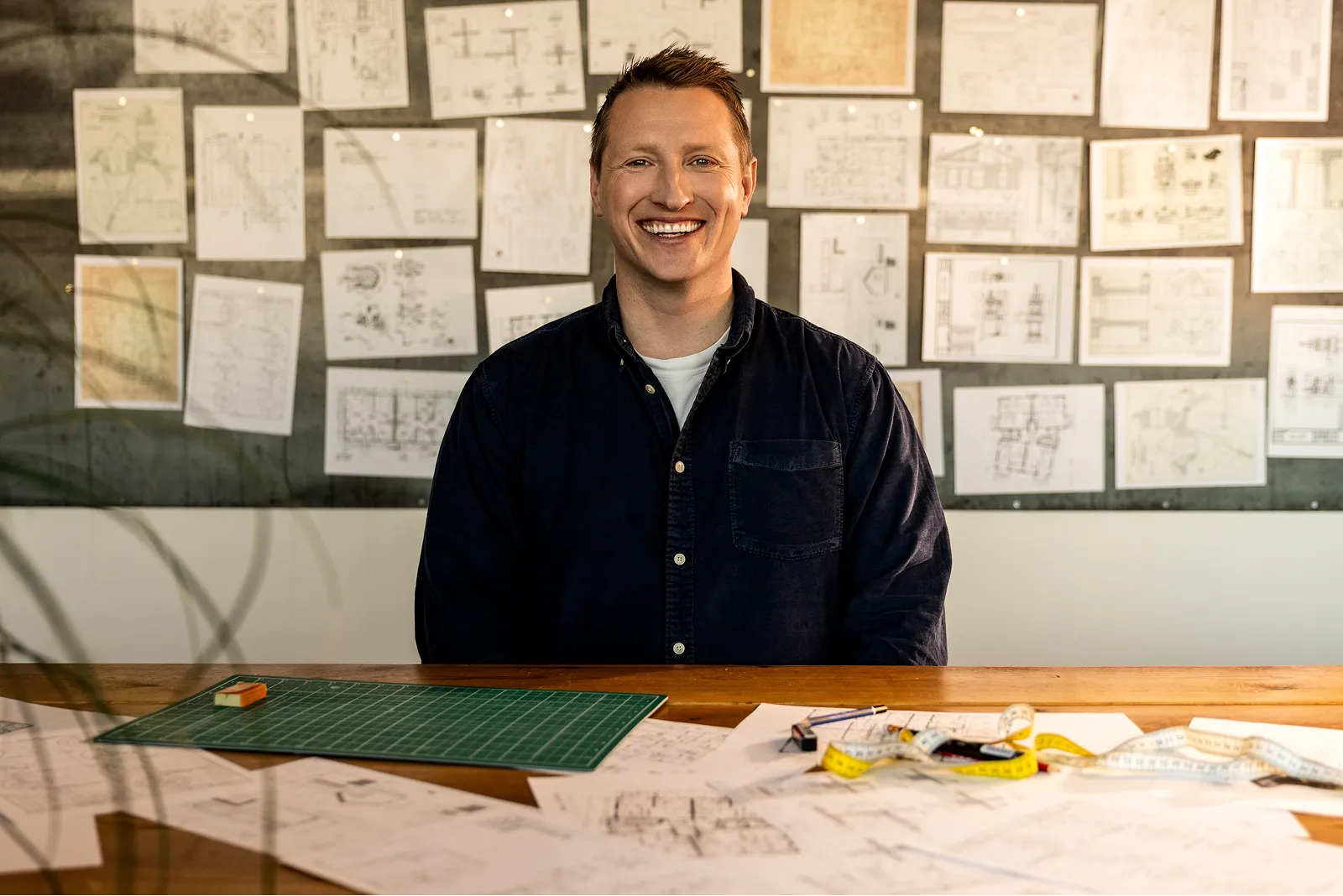 Maður í dökkri skyrtu situr brosandi framan við borð með teikningum og mælistiku, bakgrunnurinn er klæddur með arkitektúrteikningum.
