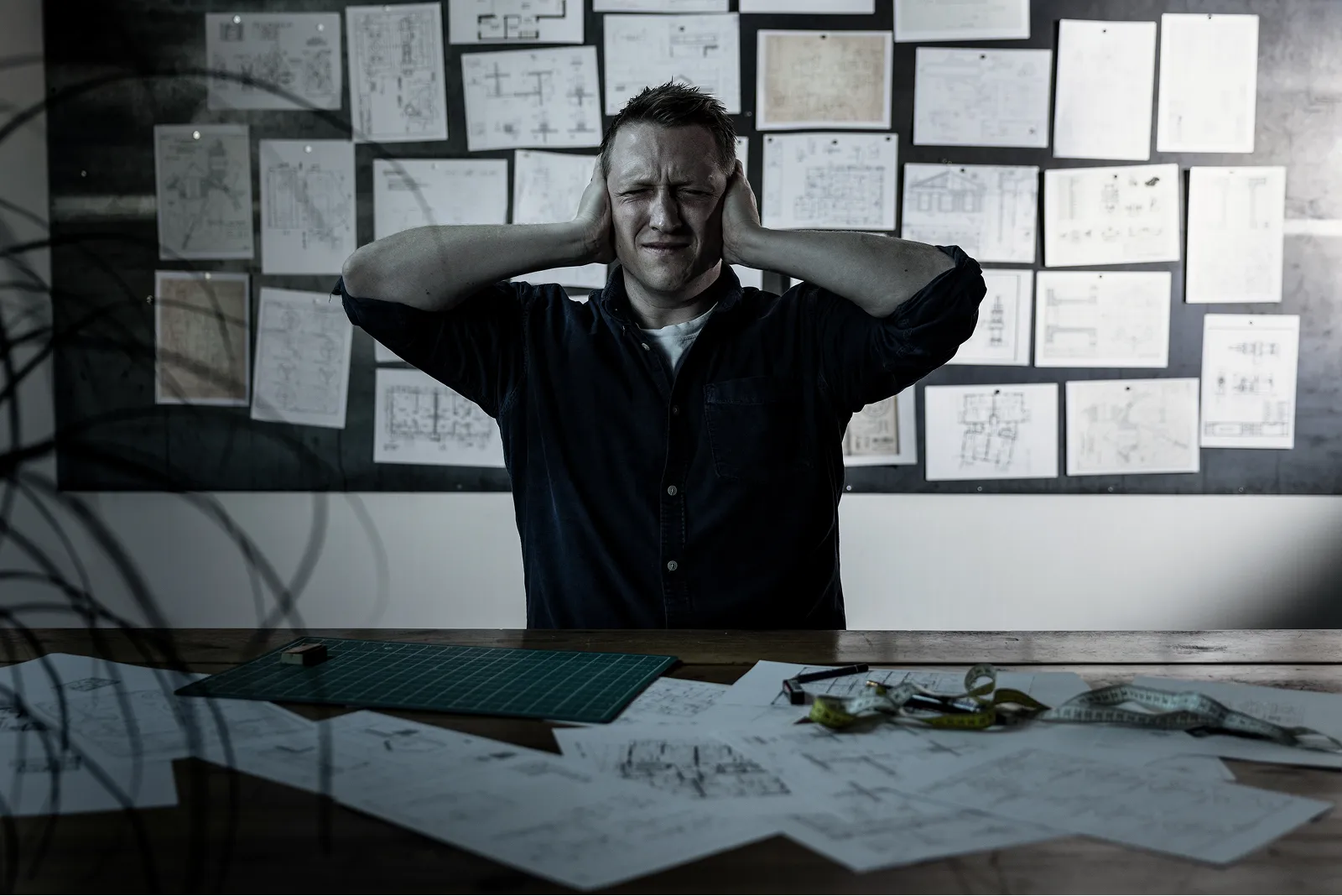 Maður situr fyrir framan hönnunaruppdrætti á vegg og borði, með höndina fyrir eyrun og andlit sem sýnir streitu.