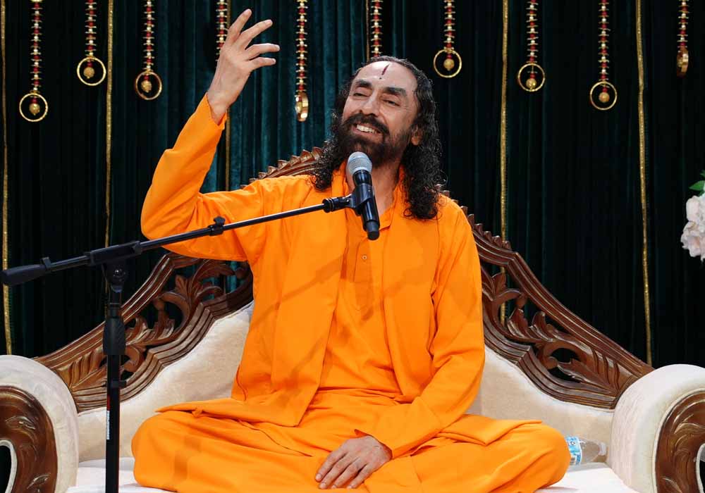 Swami Mukundananda smiling as he speaks during a satsang, seated in saffron robes with one hand raised