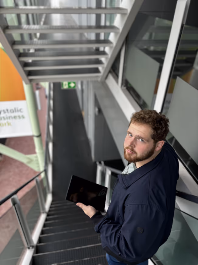Een man met een baard kijkt omhoog terwijl hij op een stalen trap in een modern kantoorgebouw staat, met een tablet in zijn hand, mogelijk bezig met werk gerelateerd aan energiebesparing of verduurzaming.