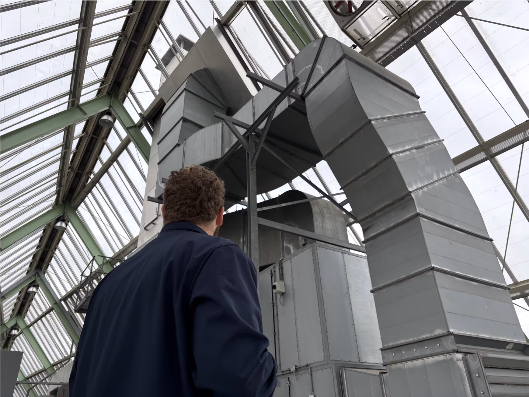 Een technicus inspecteert grote, zilverkleurige ventilatiesystemen binnen een gebouw met een glazen en metalen dakstructuur, mogelijk gerelateerd aan maatregelen voor energiebesparing of verduurzaming.