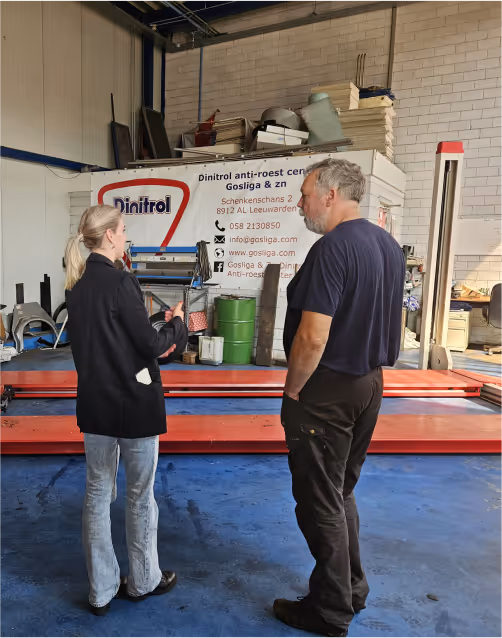 Een man en een vrouw staan in een garage en hebben een gesprek; de vrouw houdt documenten vast terwijl ze omgeven zijn door gereedschap en een groot embleem van Dinitrol, suggererend dat het gesprek over energiebesparingsmaatregelen en verduurzaming kan gaan.