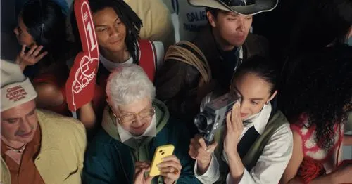 Diverse crowd at an event with an elderly woman using a yellow phone while others hold foam fingers and a camera