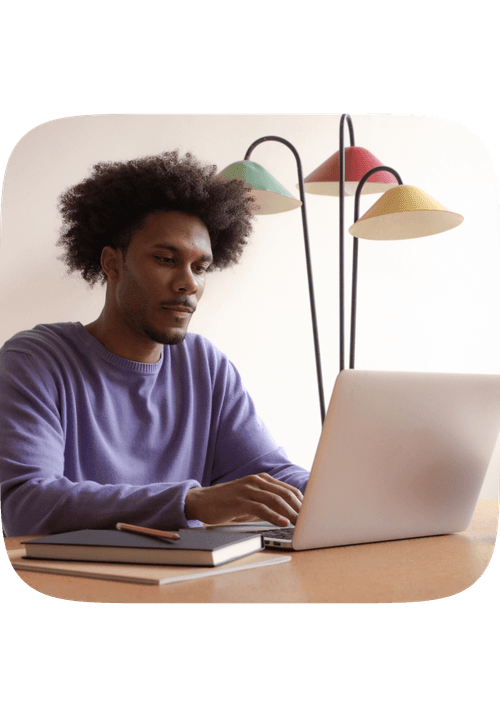 Man in purple sweater typing on laptop at desk with notebook and colorful desk lamps
