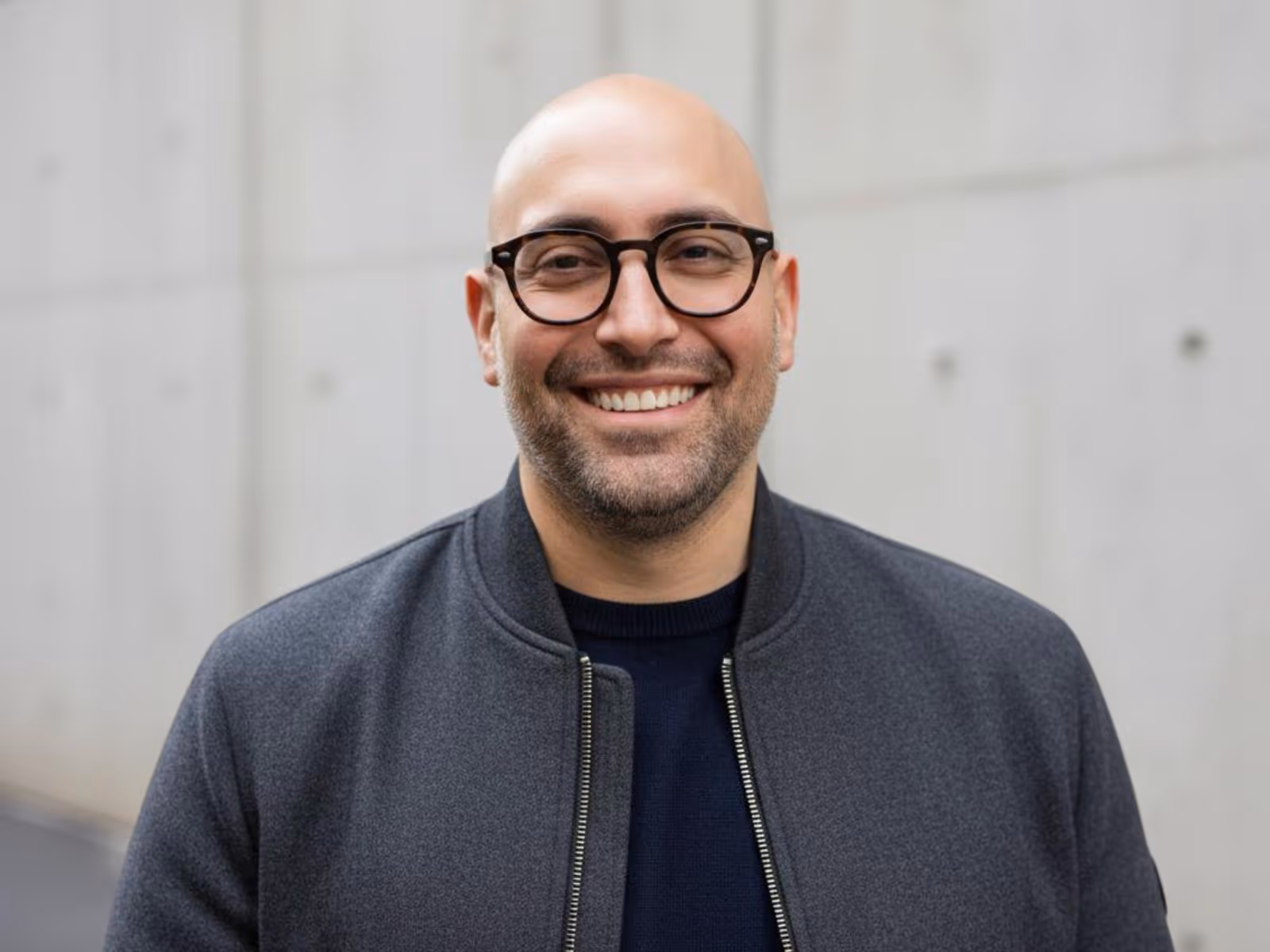 Headshot of Os Fuentes, a smiling bald man with glasses wearing a grey jacket, standing outdoors