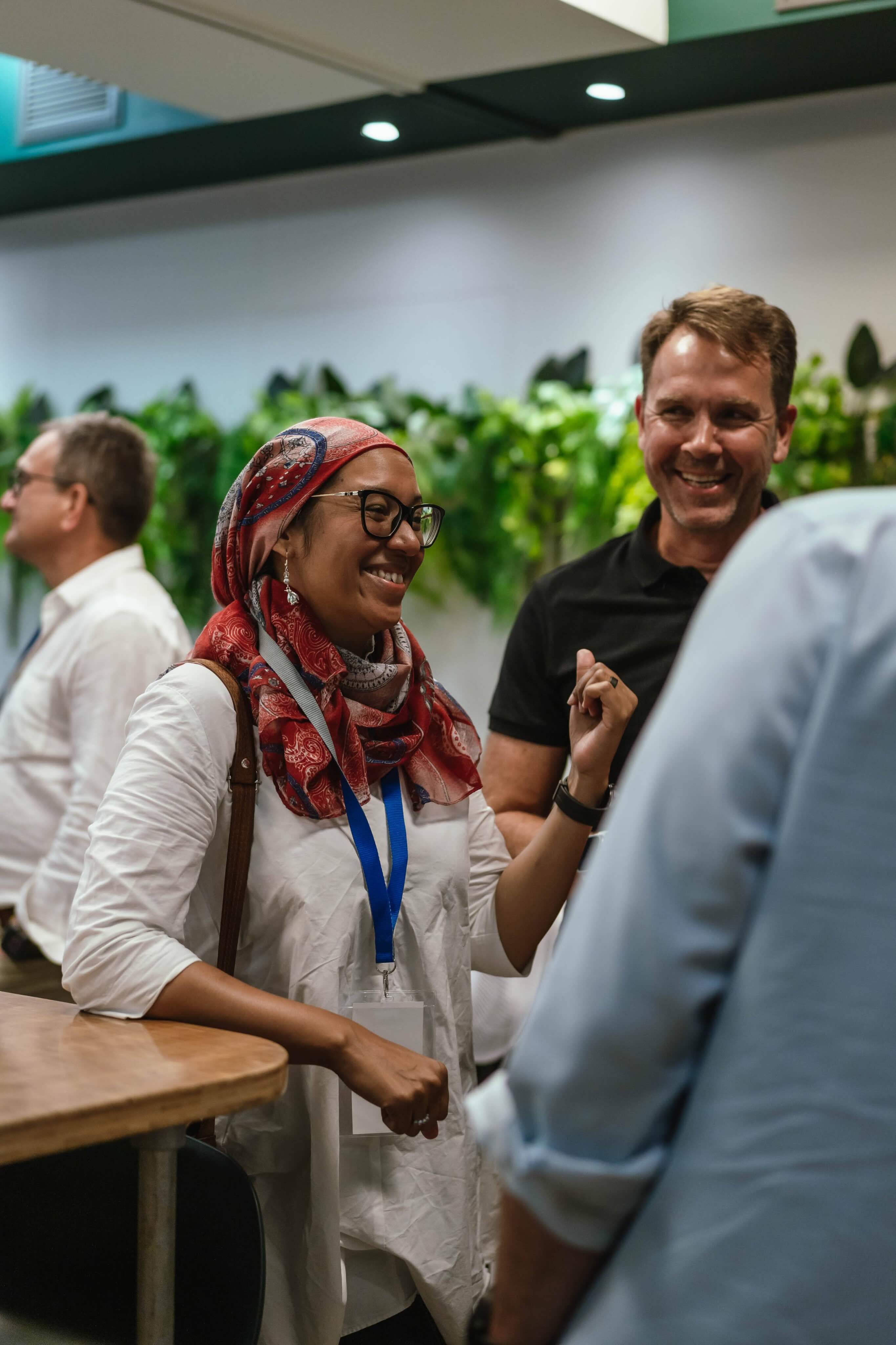 Smiling woman wearing a red patterned headscarf and glasses chatting with two men in a casual indoor setting with plants in the background.