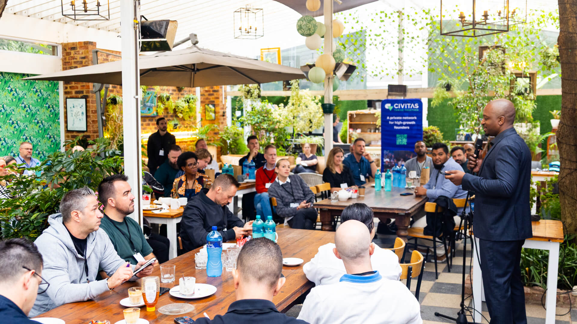 A man in a suit speaking to a diverse group seated around tables in a bright, plant-filled indoor venue with a Civitas banner in the background.