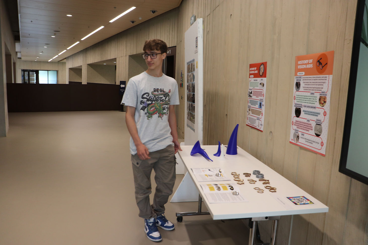 A 16 year old boy standing in a corridor next to a wall with posters on it looking sideways.