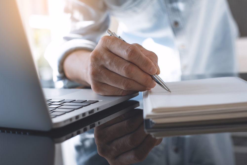 Hand taking notes with a pen next to a laptop during an expert interview