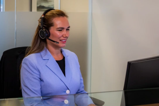 Smiling customer service representative or employee wearing a headset and a light blue blazer, working at an office desk in front of a computer monitor.