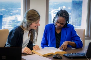 Two businesswomen engaged in office collaboration, intently reviewing a large book or case file while seated at a desk with a city skyline visible in the background.