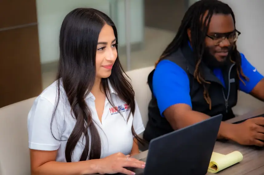 A candid shot of two colleagues, a Hispanic woman with long dark hair in a white polo shirt with a logo, and a Black man with dreadlocks and glasses in a blue shirt and black vest, working on a laptop together at a table.