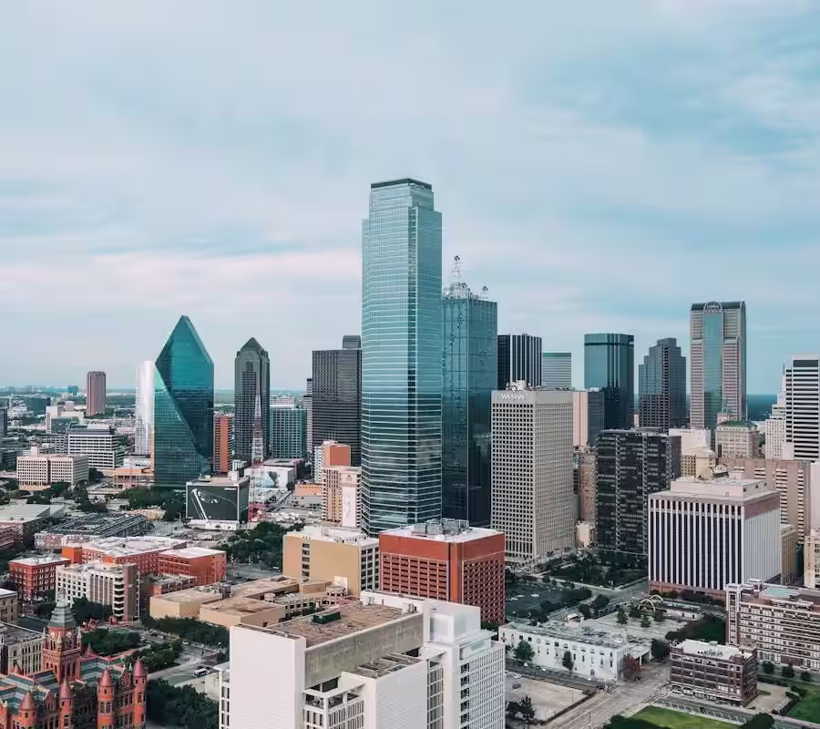 An aerial view of the Dallas, Texas, downtown skyline on a cloudy day, featuring prominent skyscrapers like the Bank of America Plaza (tallest blue reflective building) and the elongated, diamond-shaped office building (Fountain Place). Historic, shorter buildings are visible in the foreground.