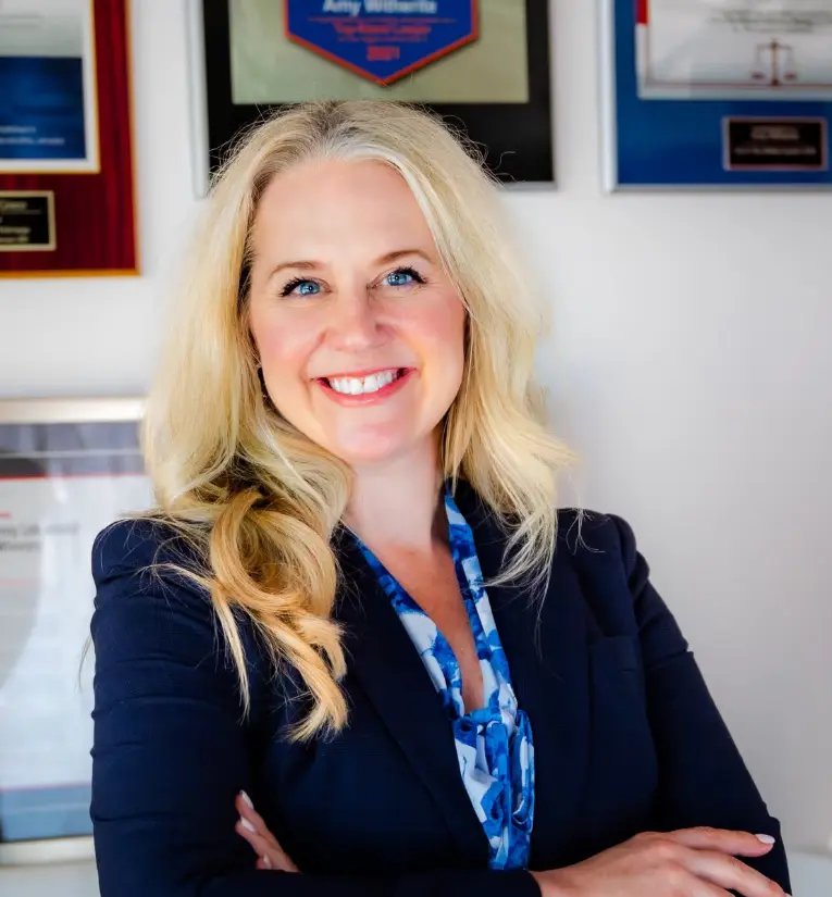 A close-up professional portrait of a smiling blonde woman with blue eyes, wearing a navy blue suit jacket and a blue and white patterned scarf. She has her arms crossed, and blurred awards plaques are visible on the wall behind her.