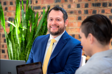 Attorney Victor in a blue suit and yellow tie smiling while meeting with a client during a consultation, with a brick wall and green plant in the background.
