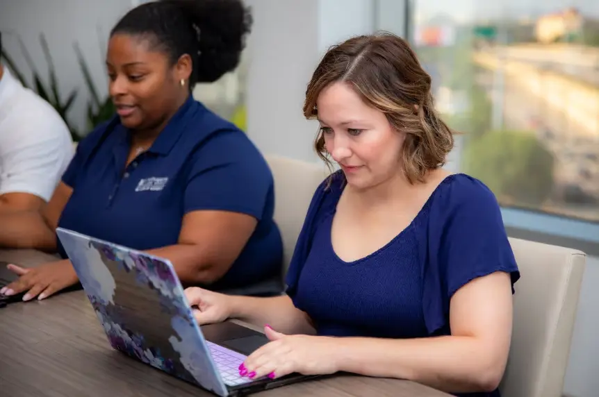 A candid shot of two women working side-by-side at a conference table in an office. The woman on the left wears a navy blue polo, and the woman on the right wears a navy blue top and is working on a laptop with a floral skin. An outdoor scene with a highway is visible through a large window.