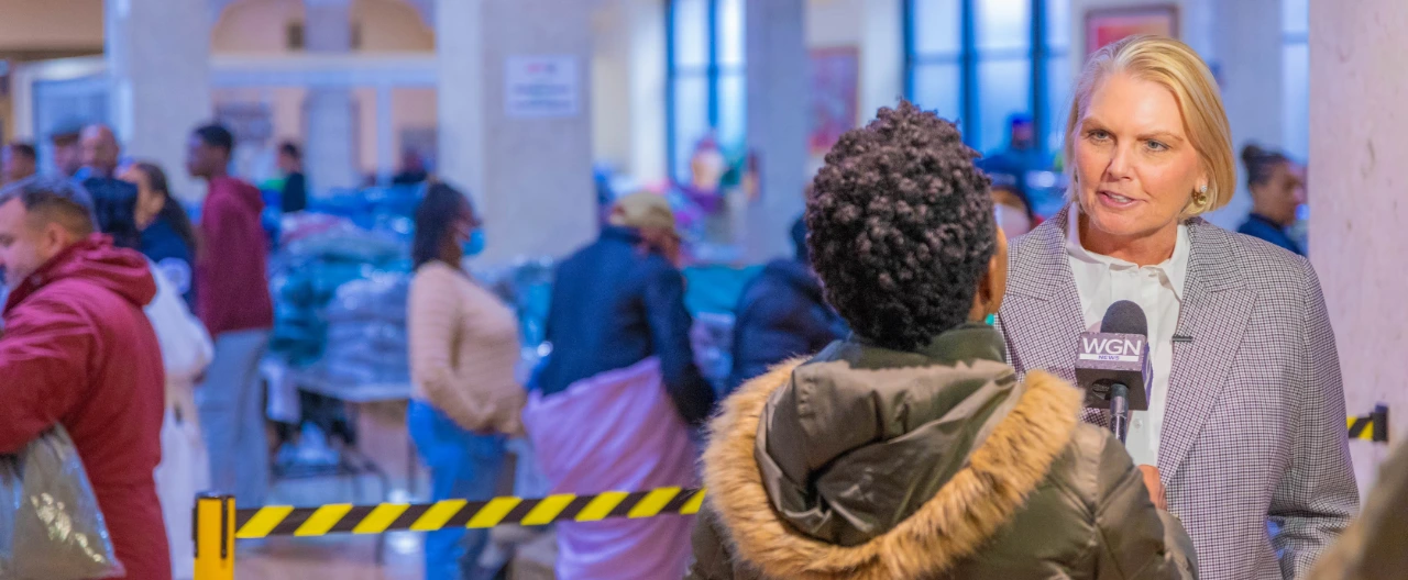 Amy Witherite giving an interview to a reporter holding a WGN branded microphone at a crowded indoor community event.