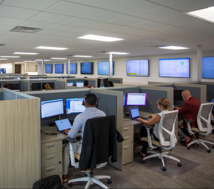 Wide shot of a busy call center or open office floor, showing employees working in cubicles with multiple monitors and several large screen data displays mounted high on the walls.