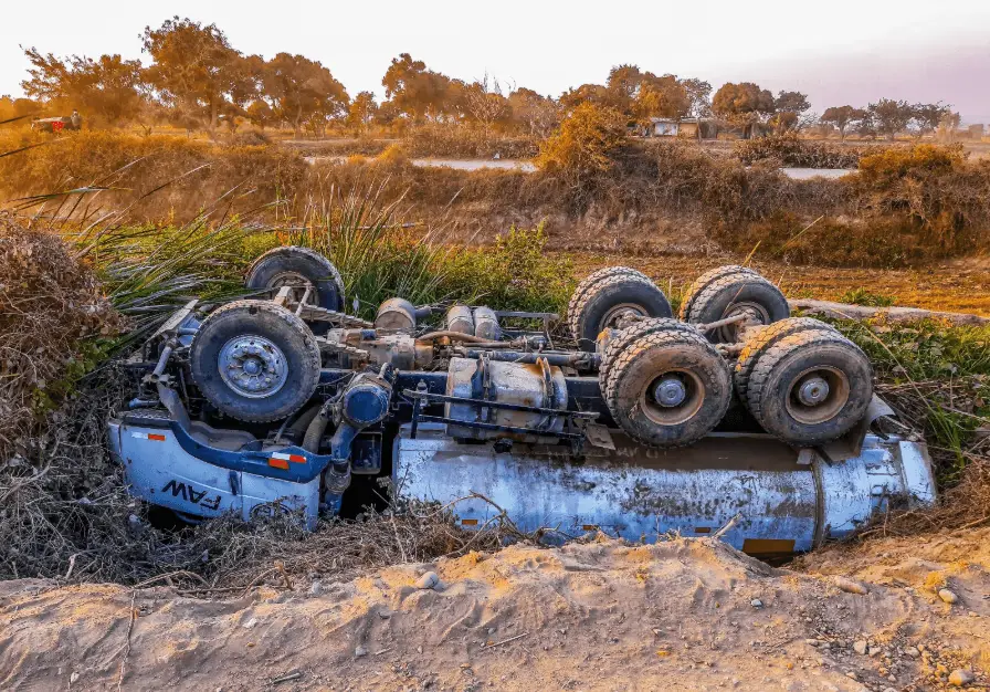 Overturned FAW tanker truck lying on its side in a ditch or embankment following an accident, showing its wheels and trailer chassis against a dry, rural background.