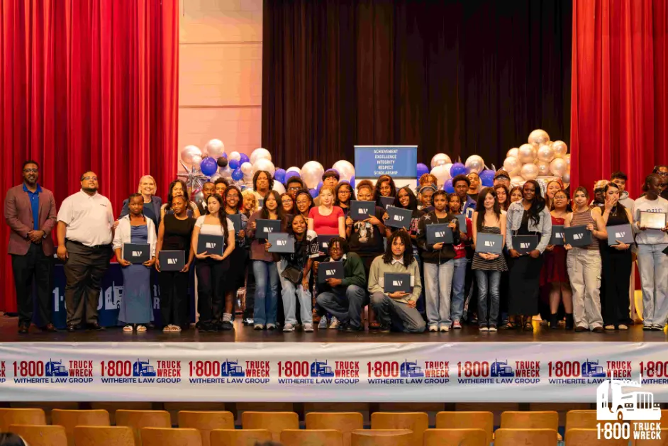 Large group photo of students and adults on a stage at a scholarship award ceremony, with students holding their diplomas or certificates, backed by red curtains and sponsored banners.