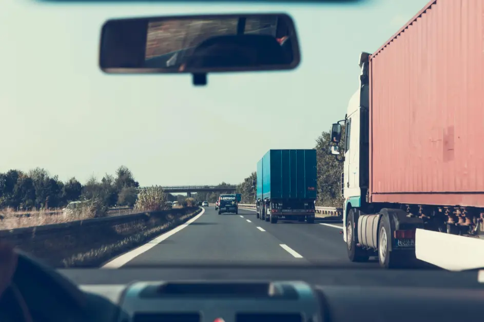 Dashboard view from a car driving on a multi-lane highway, showing two large semi-trucks with red and blue containers, and other traffic ahead.