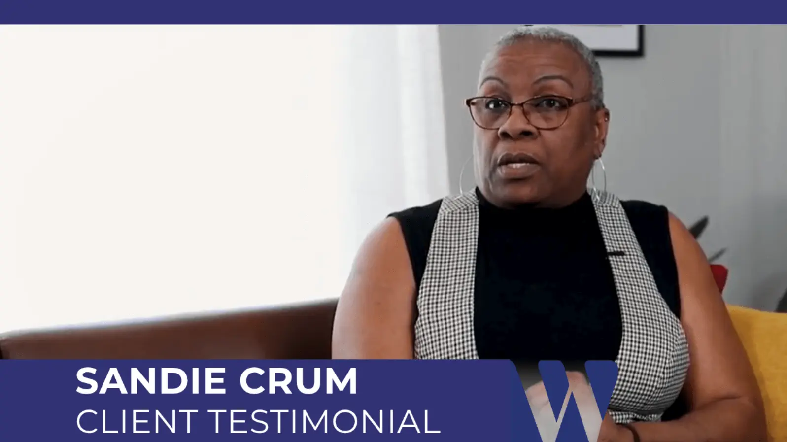 Video screenshot of Sandie Crum delivering her client testimonial. She is wearing glasses and a high-collared vest while seated indoors, with an on-screen title banner confirming the segment.