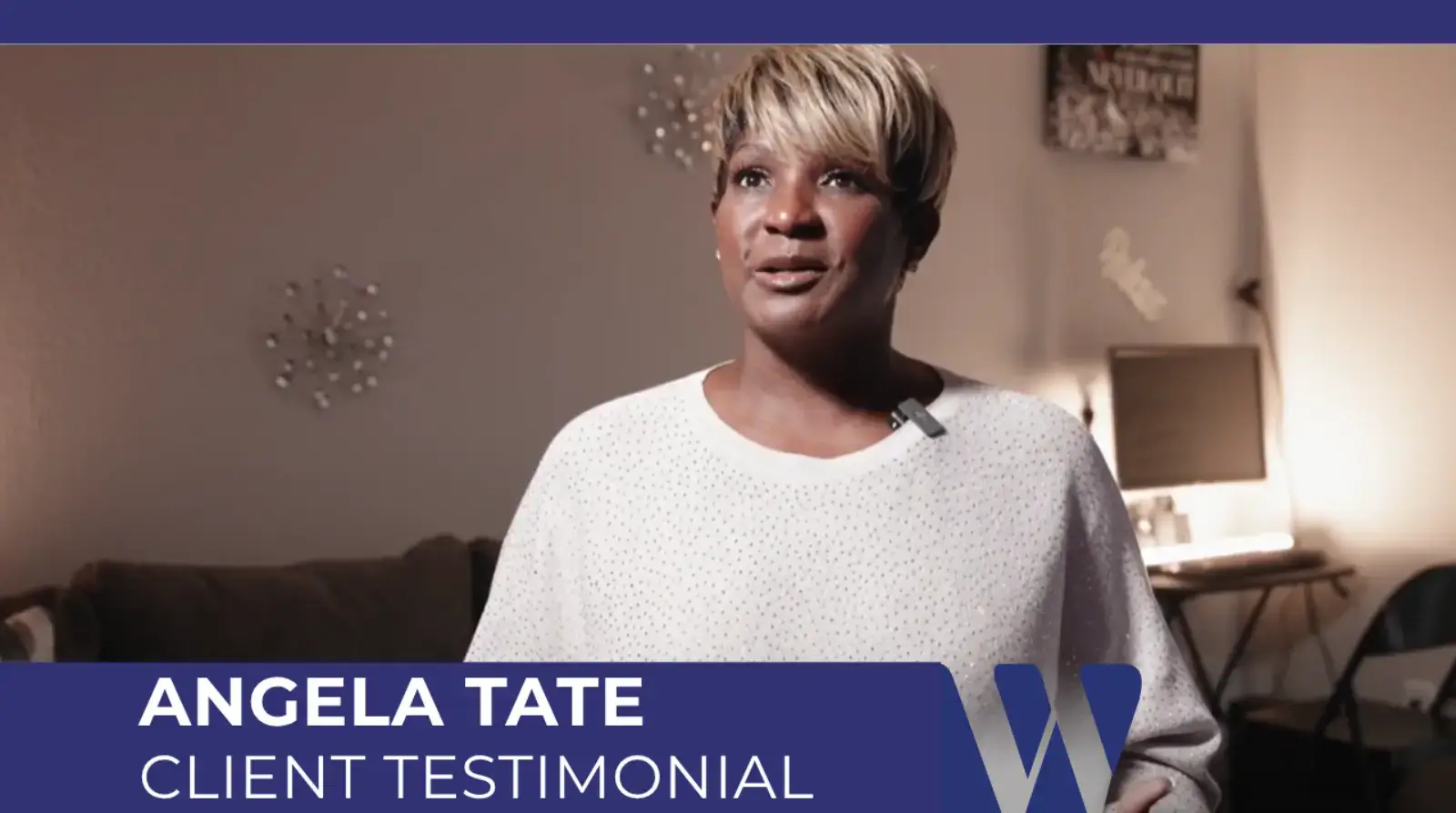 Video screenshot featuring Angela Tate giving a client testimonial indoors. She has short, blonde hair and is wearing a white sweater.