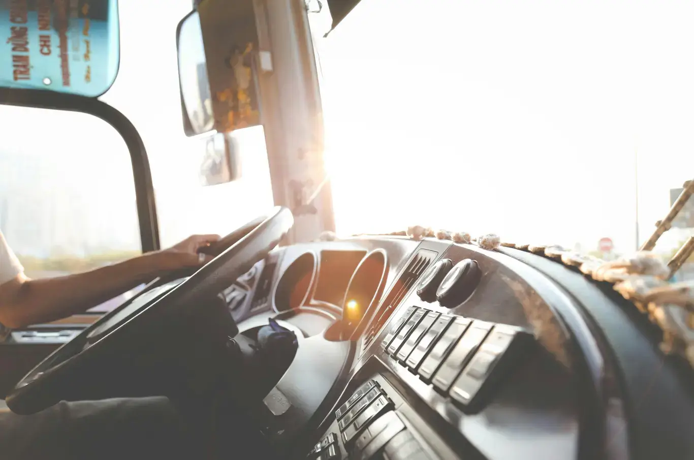 Dashboard view showing a bus driver with hands on the steering wheel, illuminated by bright sunlight streaming through the windshield.