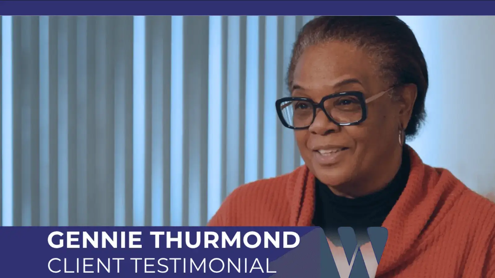 Video screenshot of Gennie Thurmond giving her client testimonial. She is wearing large glasses and an orange cardigan, seated in front of a window with blinds, and the title banner identifies her name and the segment.