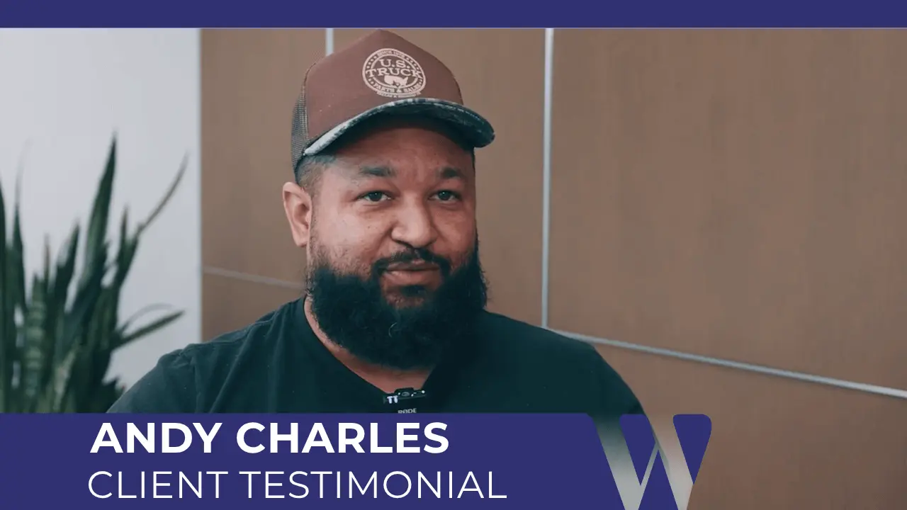 Andy Charles in a video interview, seated indoors next to a plant. He is a man with a graying beard, wearing a dark t-shirt and a dark trucker hat.