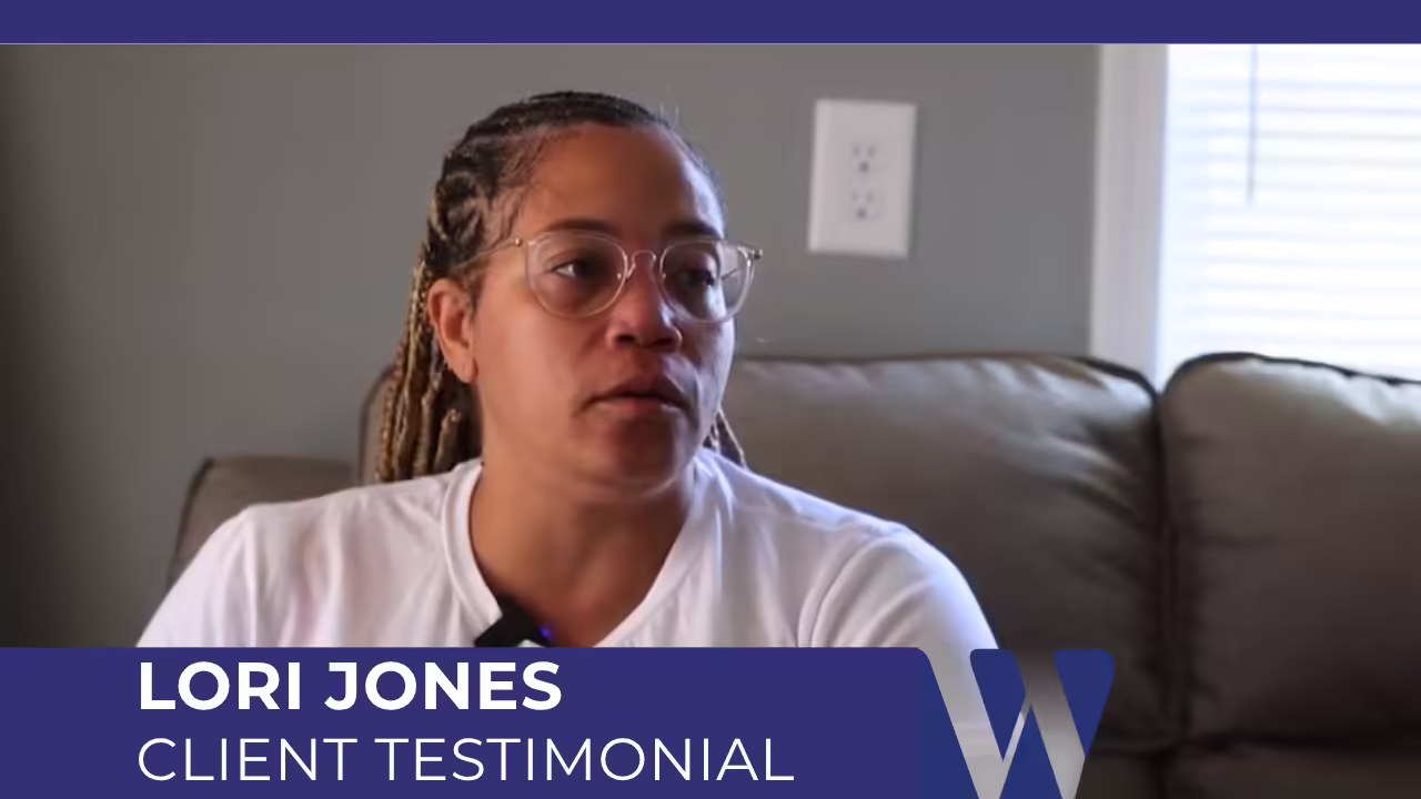 Video screenshot from an interview featuring Lori Jones giving a client testimonial. She is wearing glasses and a white shirt, with her braided hair visible, and a banner identifies the segment below.