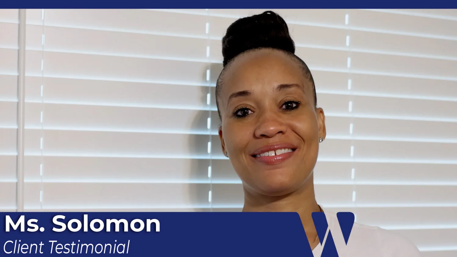 Video screenshot of Ms. Solomon giving a smiling client testimonial, wearing her hair in a high bun and seated indoors by horizontal window blinds.