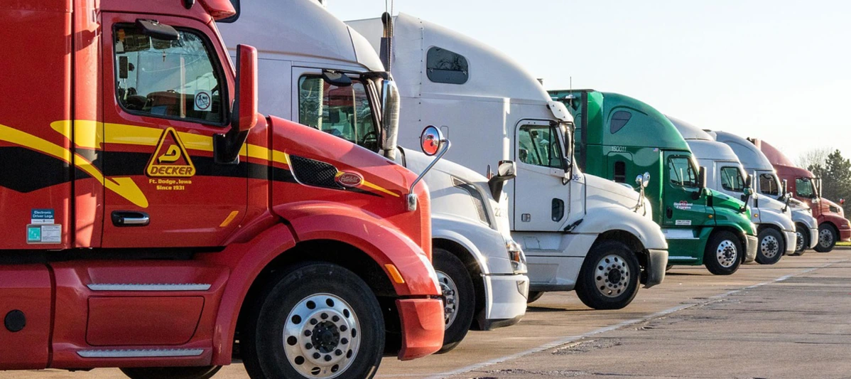 Lineup of various-colored semi-trucks (including red, white, and green) and their trailers parked closely together in a parking lot, showing a profile view of the cabs.