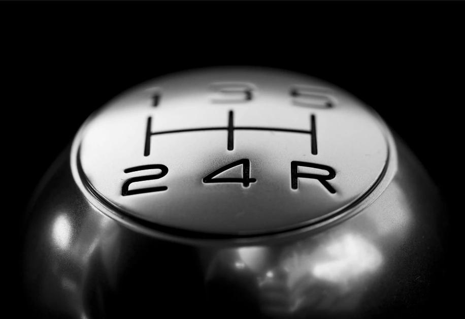 Close-up, black-and-white photo of a metallic manual transmission shift knob showing the five-speed gear pattern (1, 2, 3, 4, 5, R).