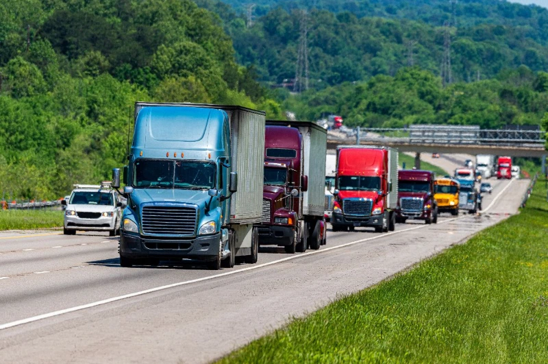 Convoy or line of semi-trucks (tractor-trailers) of various colors, including a blue lead truck, driving up a highway hill surrounded by lush green trees.