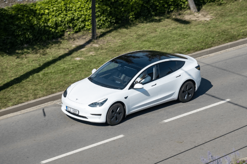 Overhead shot of a white Tesla Model 3 with a black roof driving on a paved road next to a grassy hill on a sunny day.