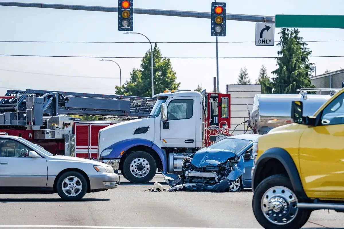 Scene of a semi-truck accident at a signaled intersection, showing a badly damaged blue car crushed between the white semi-truck and other vehicles, with emergency fire equipment visible in the background under traffic lights.