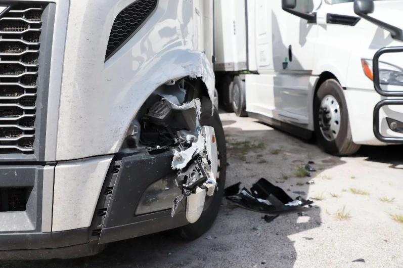 Close-up view of damage to the front fender and missing headlight of a gray semi-truck after an accident, with debris on the ground and another truck visible in the background.