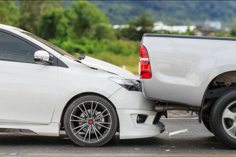 Close-up of a rear-end collision where a white car has crashed into the back of a silver pickup truck, resulting in front-end damage to the car.