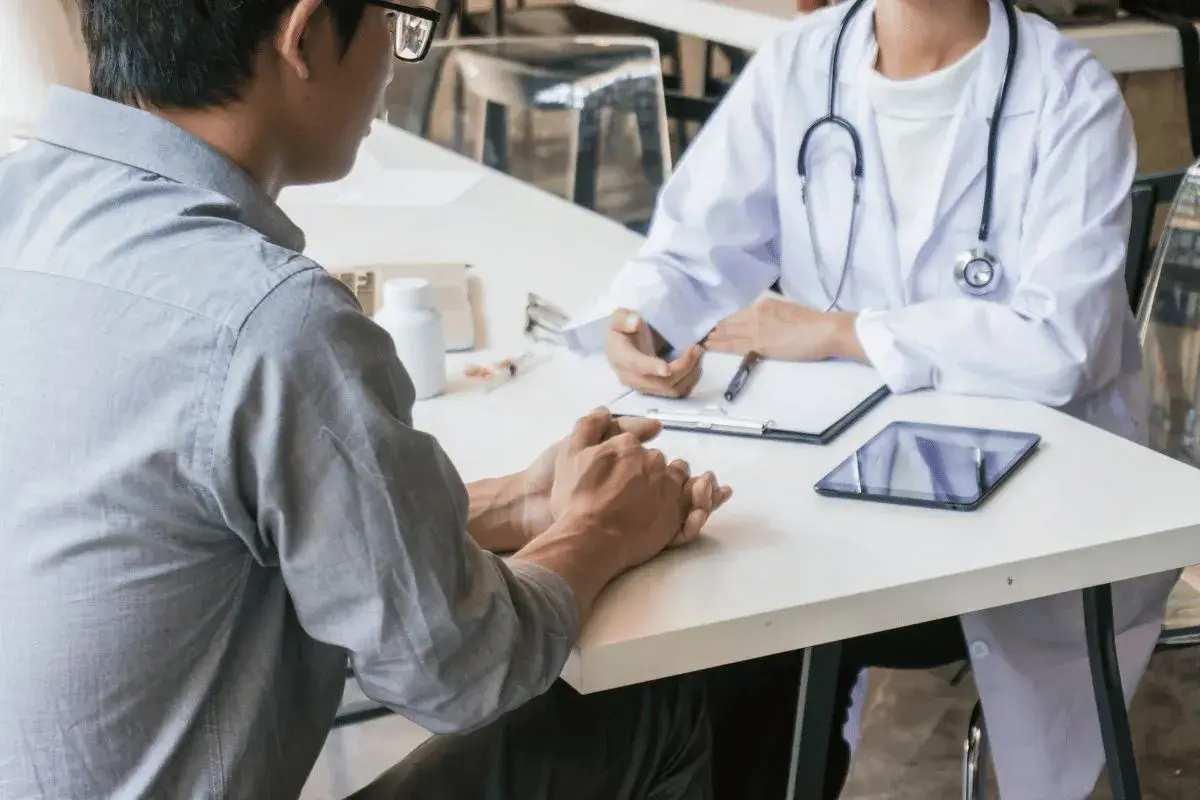 Doctor (wearing a lab coat and stethoscope) having a serious injury discussion with a patient across a table that holds a clipboard and tablet.