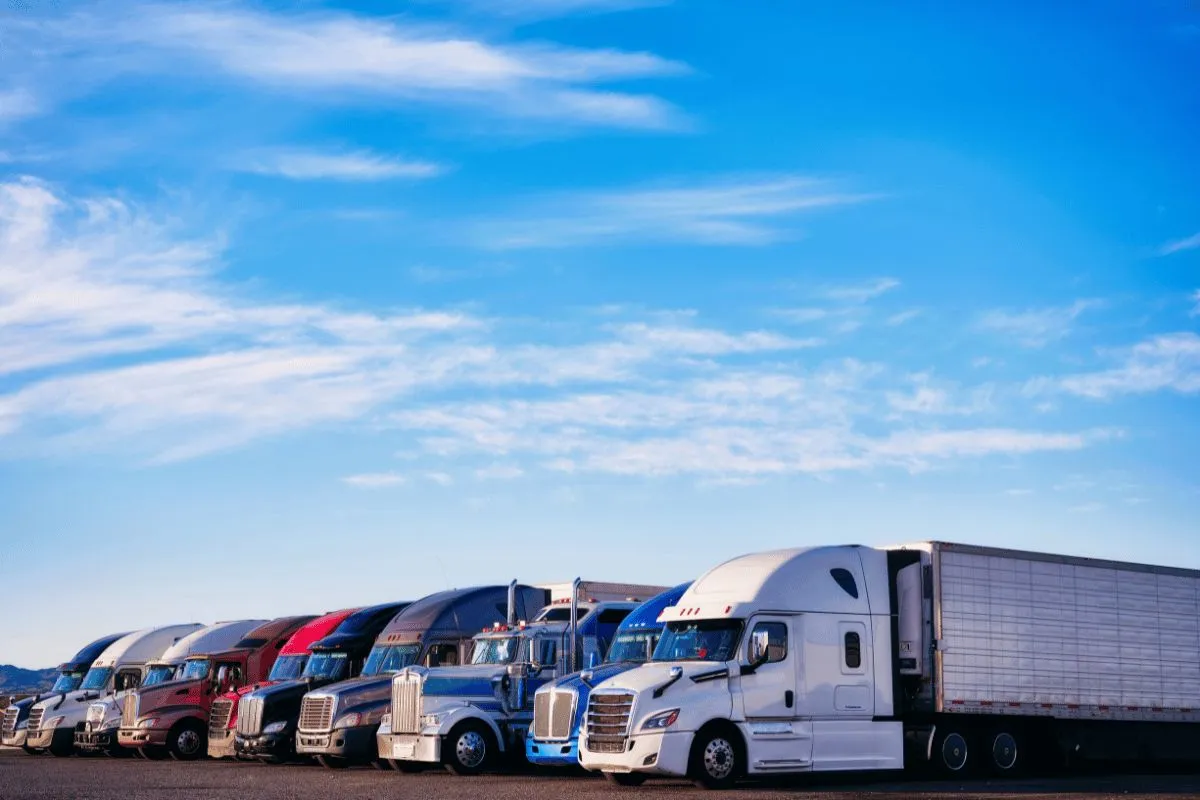 A lineup of semi-trucks (tractor-trailers) of various colors parked side-by-side at a truck depot or lot, set against a wide blue sky with light clouds.