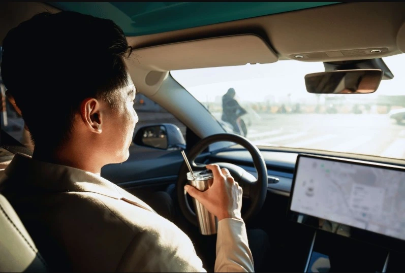 Man driving a modern car (likely an EV), seen from a dashboard view, drinking from a sustainable cup while the large central screen displays a map.