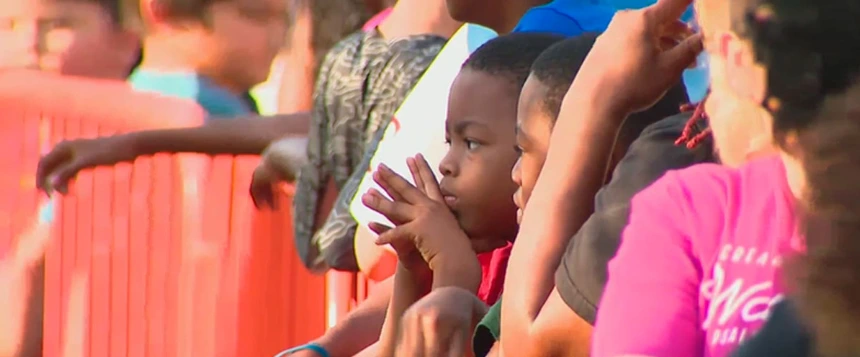 A close-up shot of two young Black boys watching an event, with their faces near an orange protective barrier or fence. The boy in the foreground has his hands clasped near his face, looking intently off-camera.
