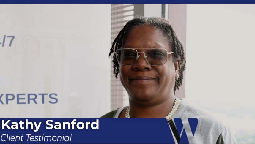A middle-aged Black woman, identified as Kathy Sanford in a lower graphic, smiles at the camera. She has dreadlocks and is wearing glasses and a chain necklace.