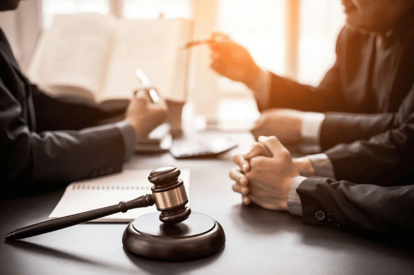 A wooden gavel rests on a conference table in the foreground, with three people in suits—likely lawyers or a legal team—seated around the table in the background. One person is reading from a large book, suggesting a legal meeting or consultation.